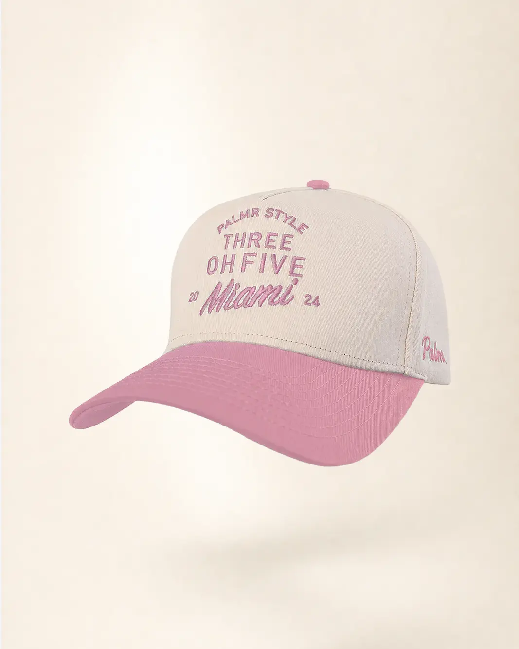 A baseball cap with a light beige crown and a dusty pink brim, featuring embroidered pink text on the front and side.