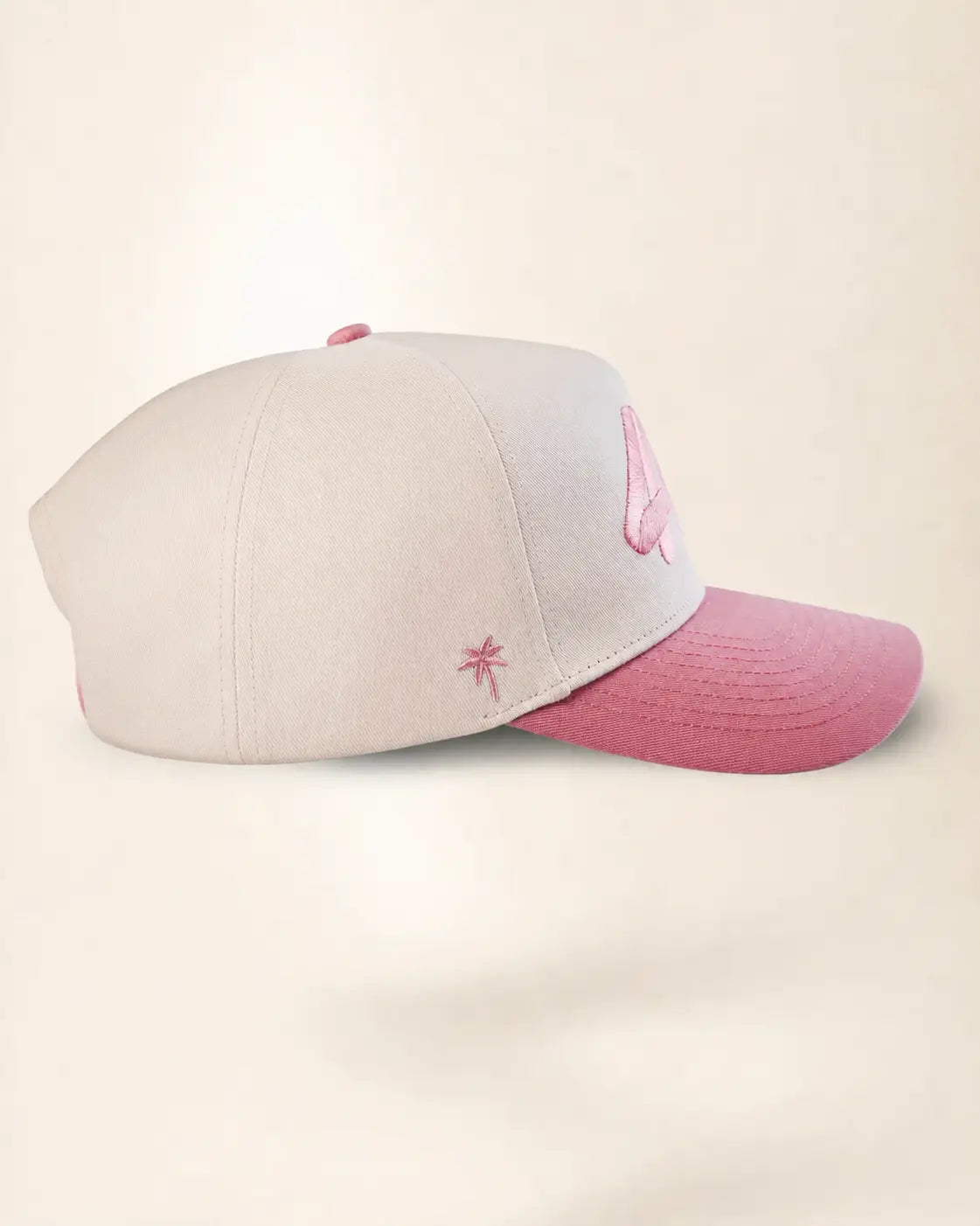 A baseball cap with a light gray crown and a dusty pink brim, featuring a small pink embroidered palm tree on the side and a pink embroidered letter on the front.