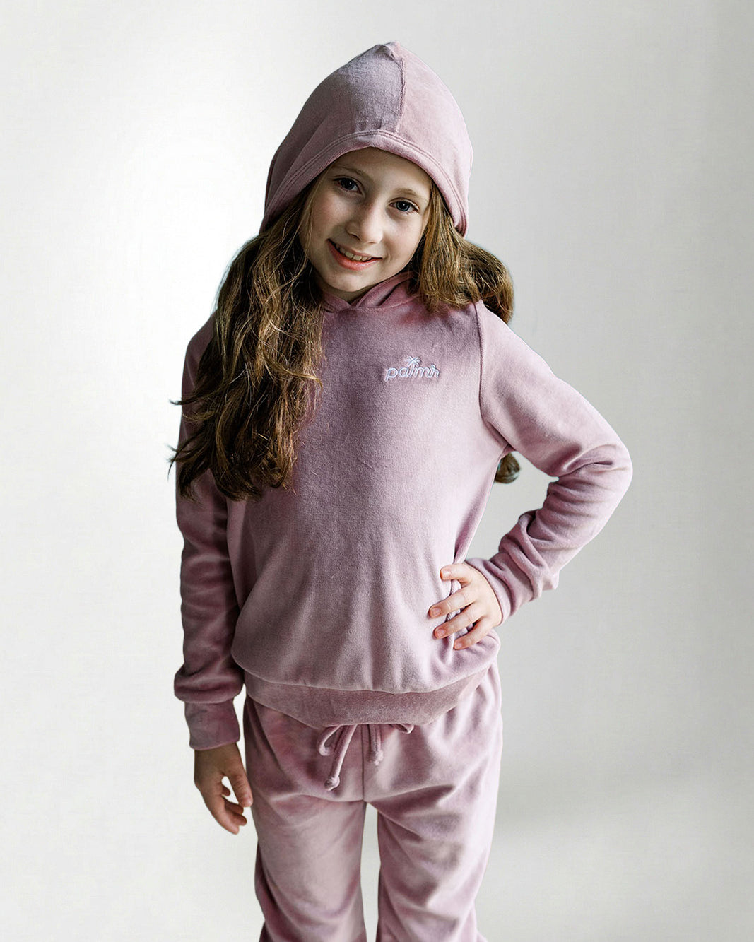 Young girl wearing a pink hoodie and pants set with a logo on a light gray background
