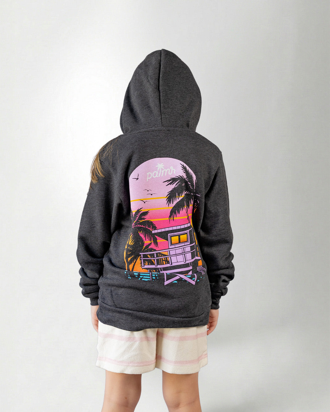 Person wearing a hoodie with a sunset and palm tree design on the back, standing against a plain background.