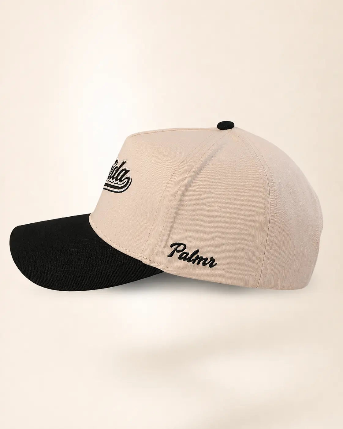 A structured baseball cap with a light beige crown and a black curved brim, featuring black embroidered script logos on the side, set against a plain cream background.