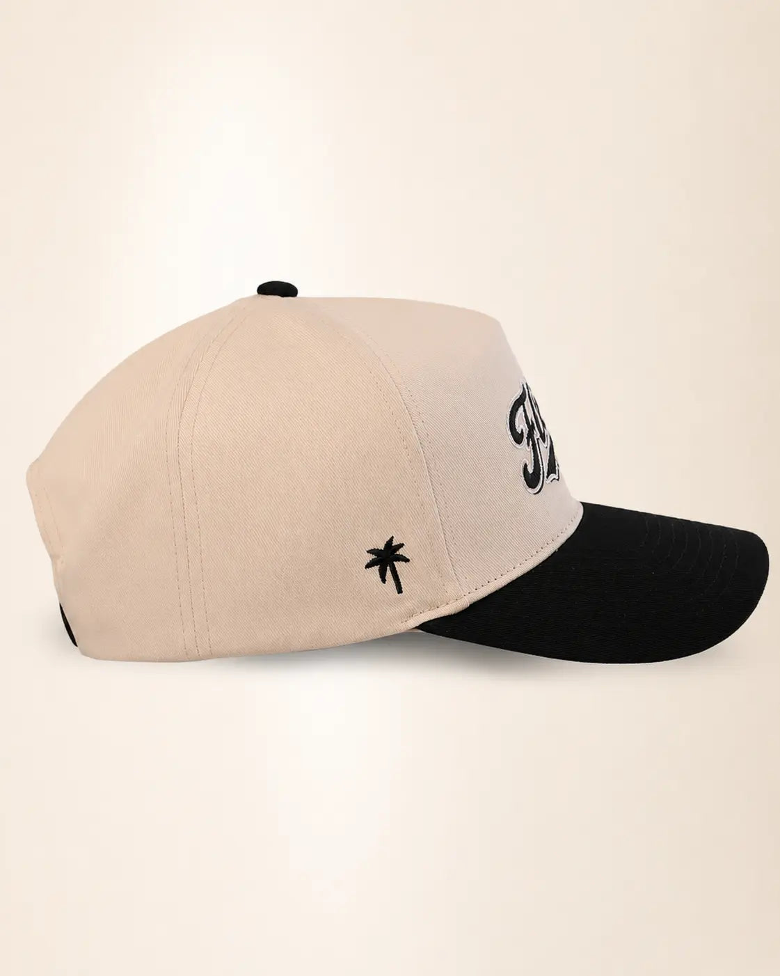 A two-tone baseball cap with a light beige crown and a black curved brim, featuring a small black palm tree logo on the side and a large black embroidered monogram on the front.