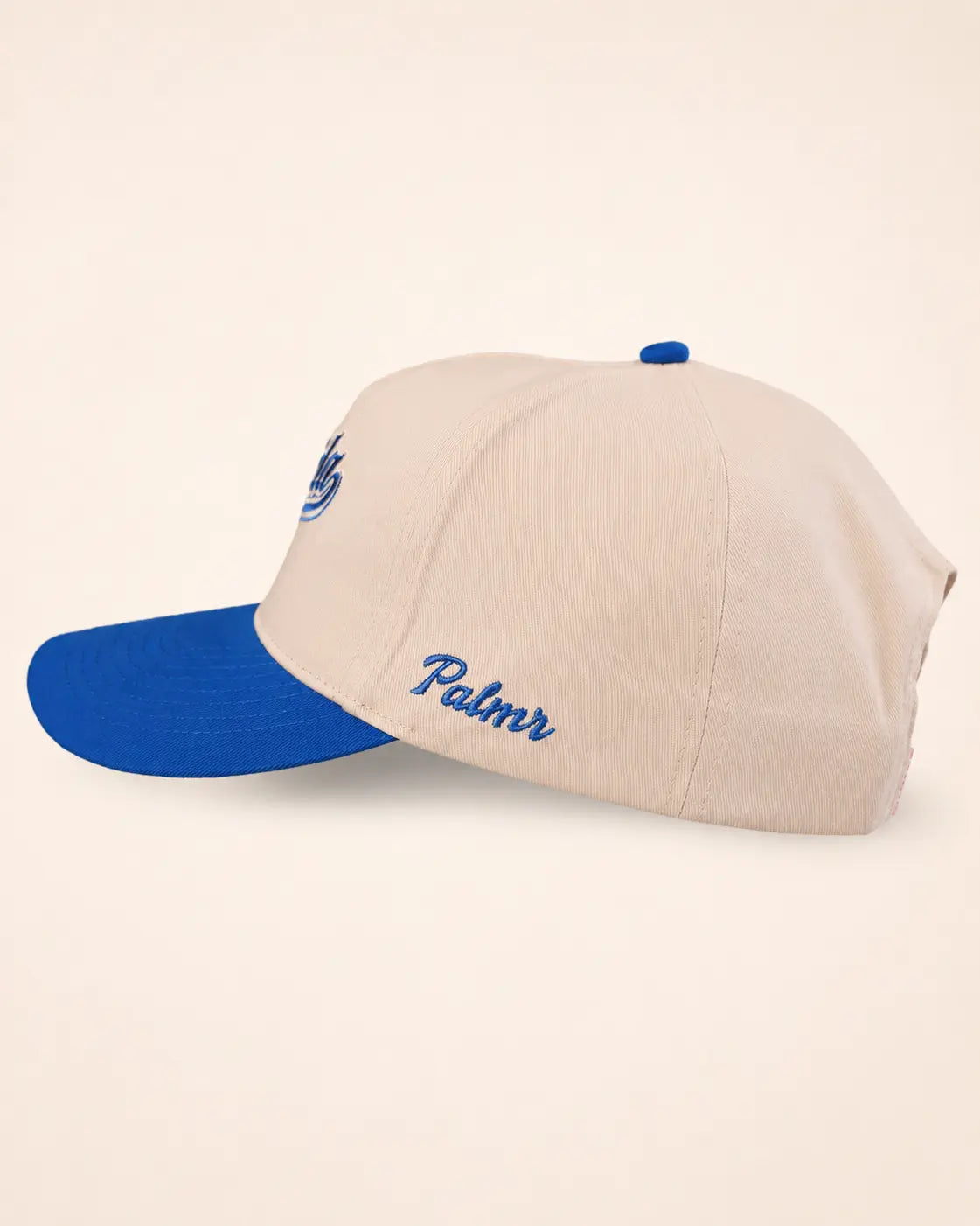 A structured baseball cap in beige with a bright blue visor, blue button, and blue embroidered script on the side, photographed on a plain light beige background.