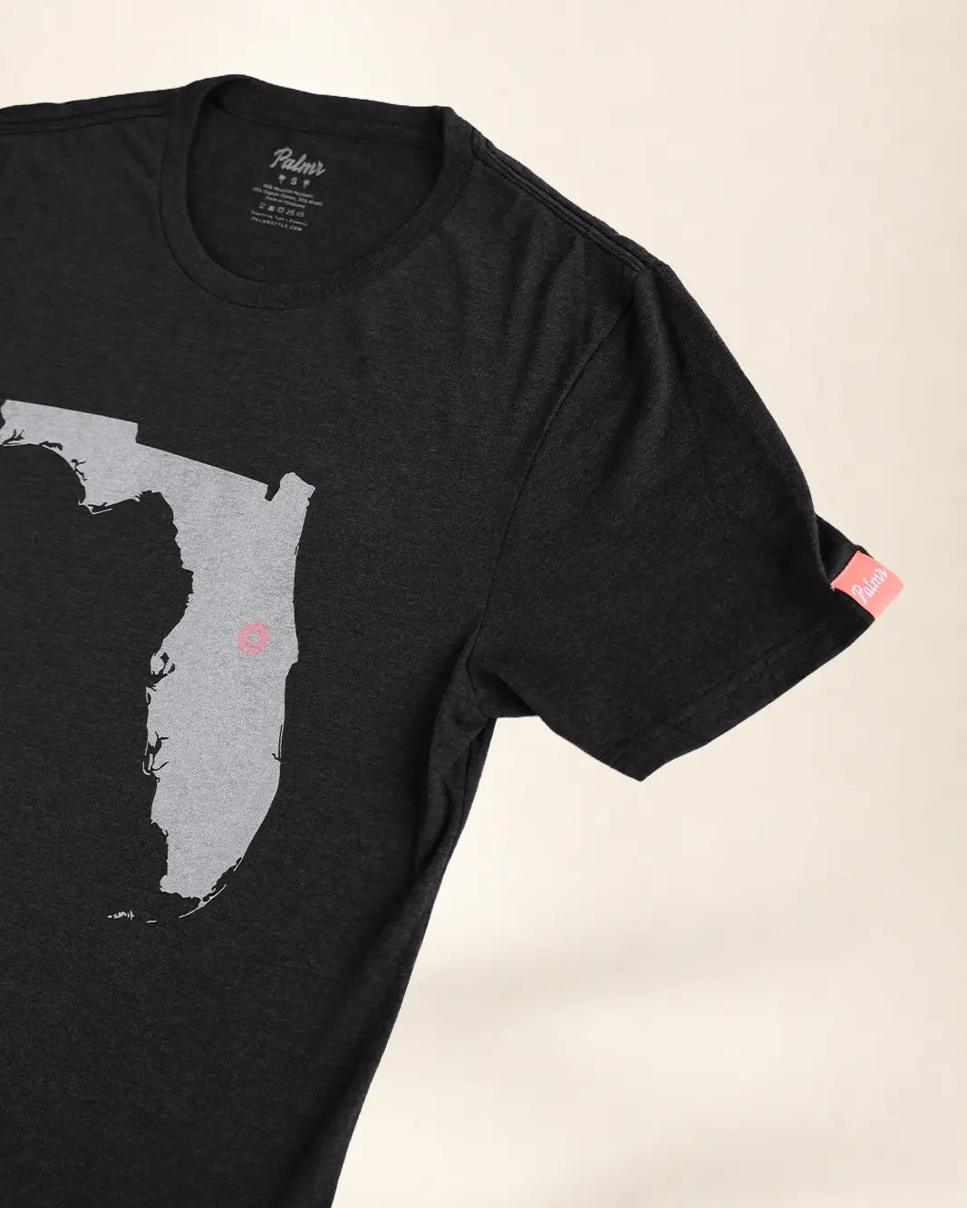A close-up of a black short-sleeve t-shirt featuring a gray silhouette map of Florida with a small Palmr logo on the interior, shown against a light beige background.
