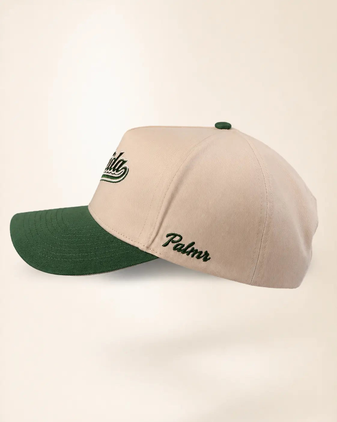 A beige baseball cap with a dark green visor and green button, featuring embroidered script logos on the front and side.