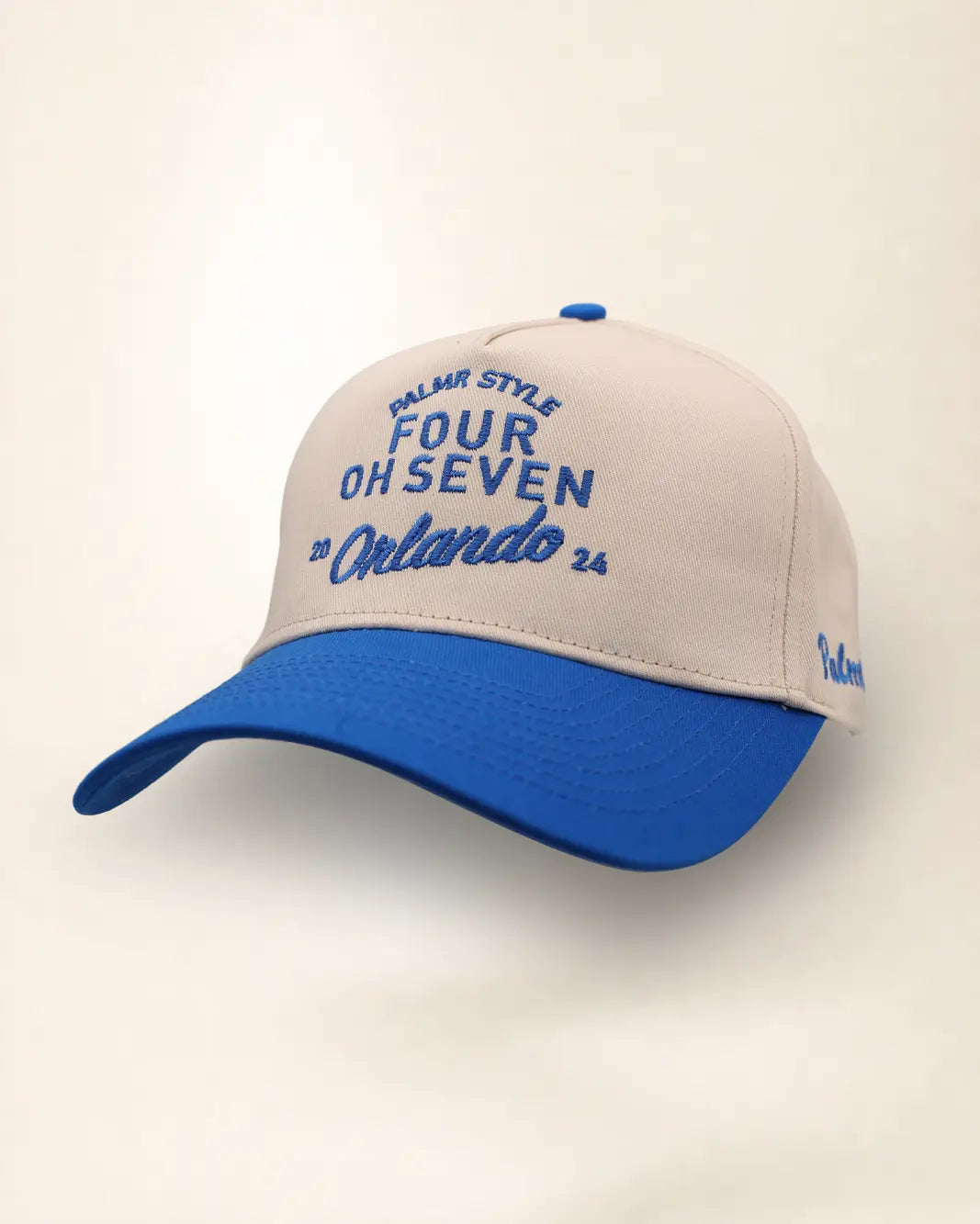 A two-tone baseball cap with a cream-colored front panel and brim, and a royal blue curved brim and top button. The front panel features blue embroidered text reading 'PALMR STYLE', 'FOUR OH SEVEN', and 'Orlando 26'.