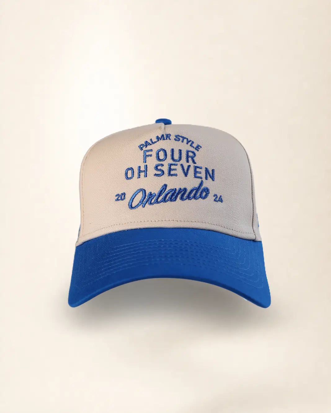 A two-tone baseball cap with a cream-colored front panel and brim, and a royal blue curved brim and top button. The front panel features blue embroidered text reading 'PALMR STYLE', 'FOUR OH SEVEN', and 'Orlando 26'.