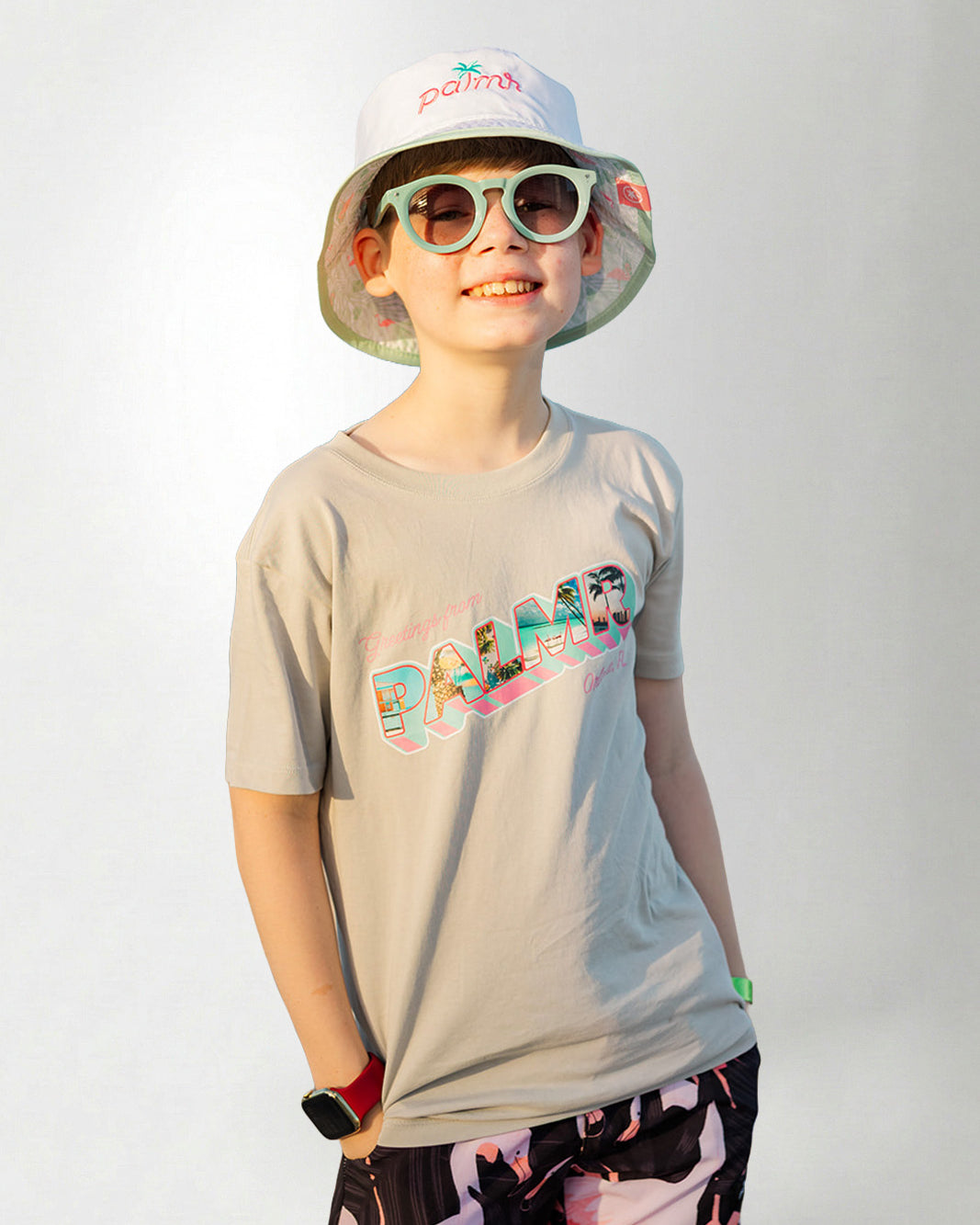 Person wearing a light gray t-shirt with colorful text, black shorts with pink designs, and a white bucket hat with green rim on a plain background.
