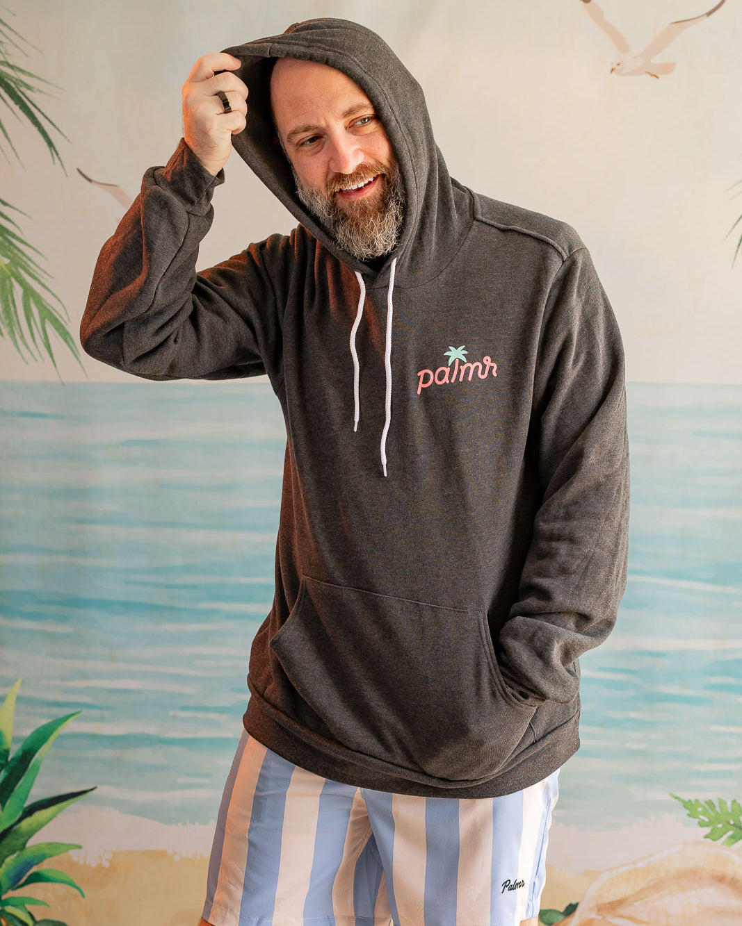 Man wearing a hoodie with a palm tree design in front of a beach mural.