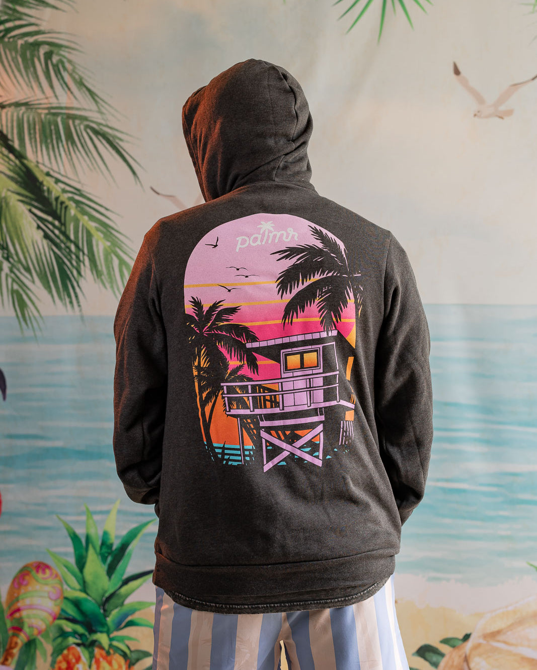 Person wearing a hoodie with a palm tree and beach house design, standing in front of a tropical-themed wall.