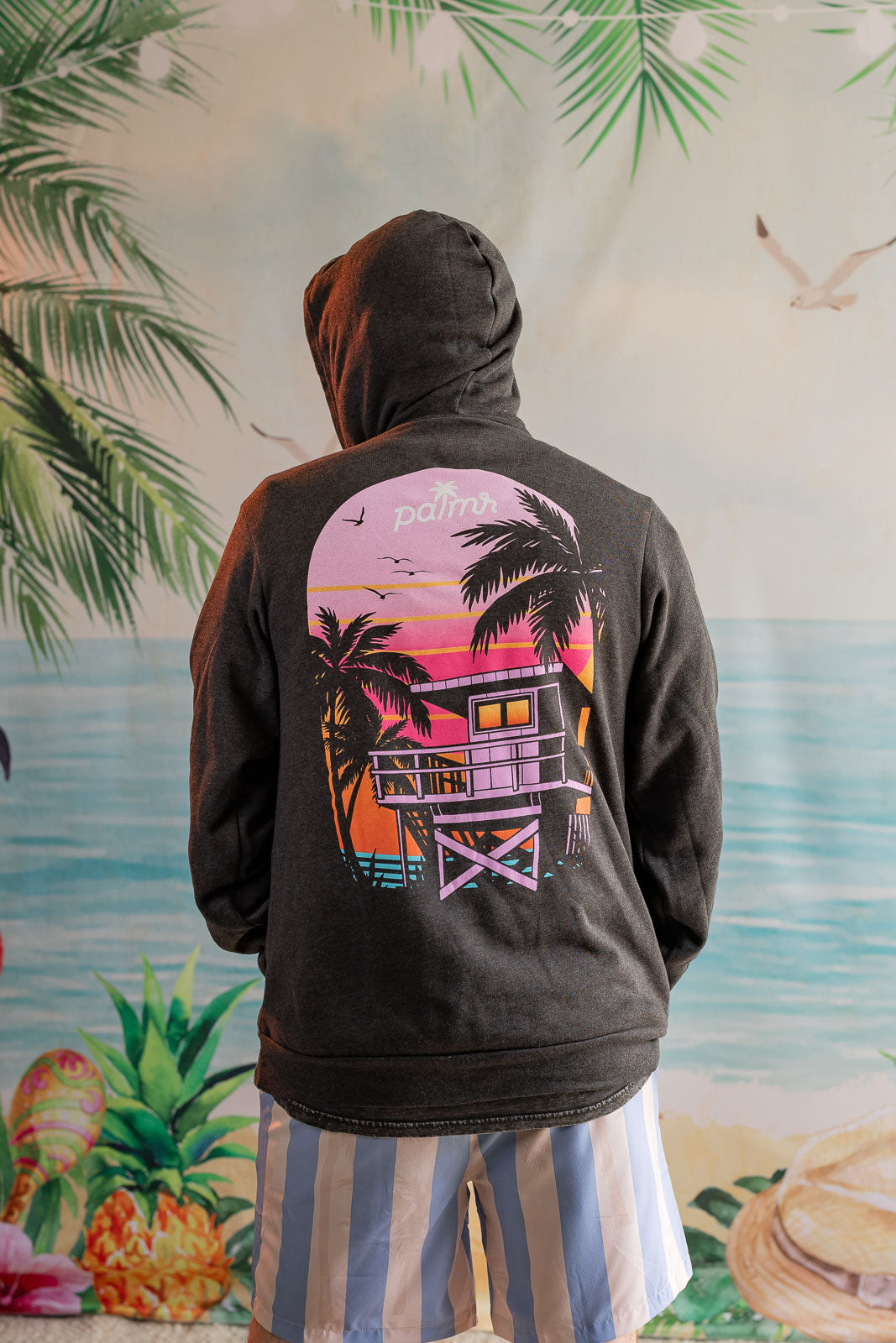 Person wearing a hoodie with a palm tree and beach house design, standing in front of a tropical-themed wall.