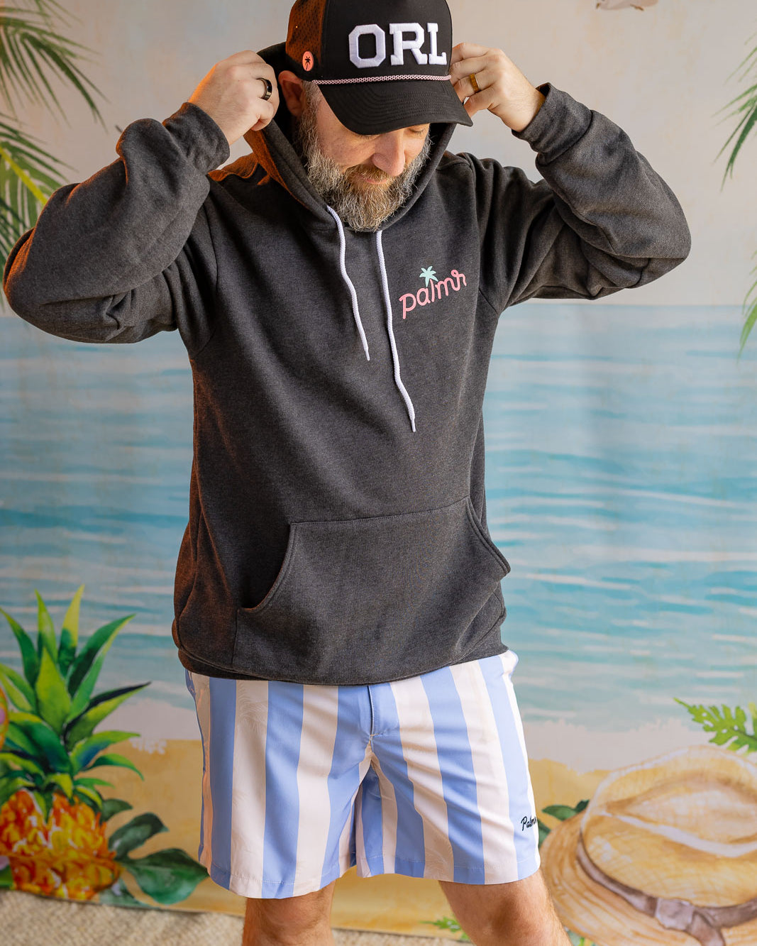 Man wearing a black hoodie with 'ORL' cap and striped shorts against a beach-themed backdrop