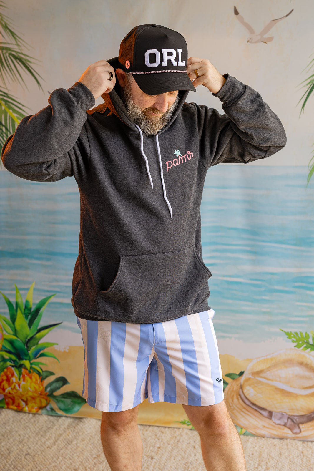 Man wearing a black hoodie with 'ORL' cap and striped shorts against a beach-themed backdrop