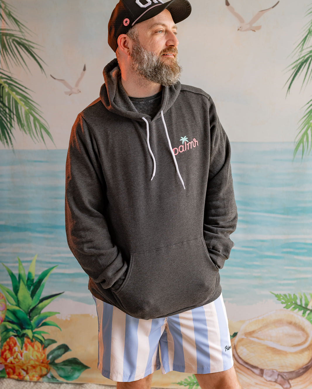 Man wearing a dark hoodie with a logo and striped shorts standing in front of a beach-themed wall mural.