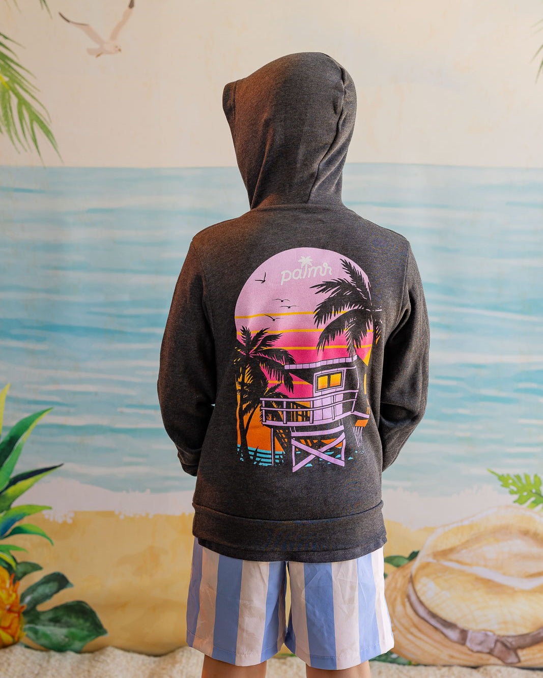 Person wearing a hoodie with a colorful lifeguard tower design in front of a beach mural.