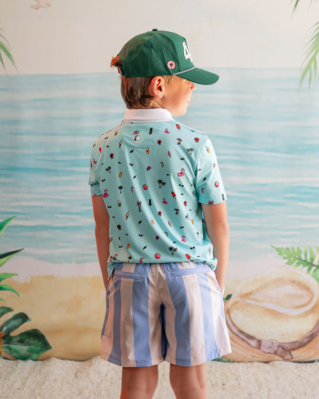 Child wearing a patterned shirt and cap, standing against a beach-themed wall.