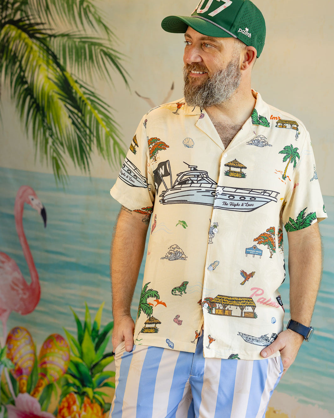 Man wearing a tropical-themed shirt and cap with a colorful background featuring flamingos and palm trees.