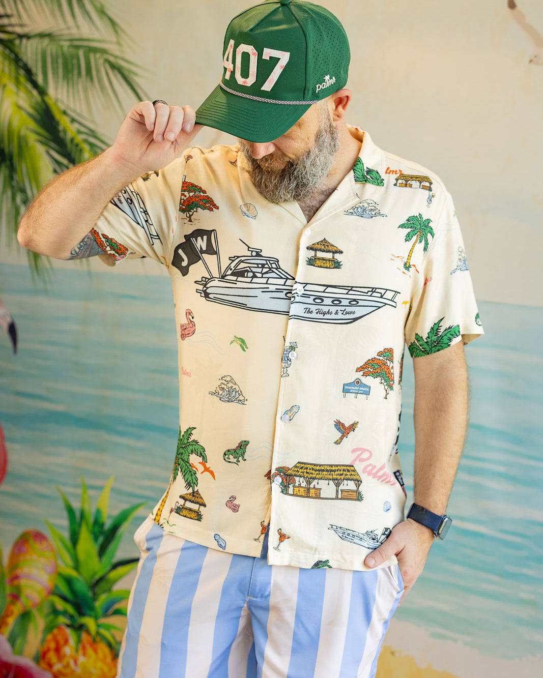 Man wearing a patterned shirt and striped shorts with a green cap, standing against a tropical-themed background.