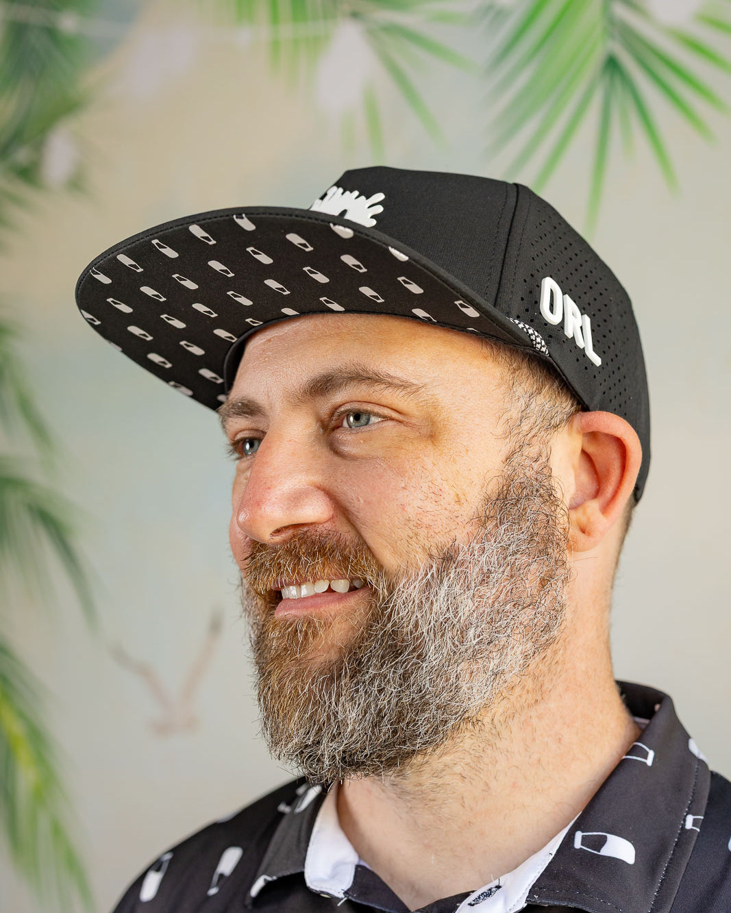Man wearing a black cap with white patterns and text, standing against a blurred green leafy background.