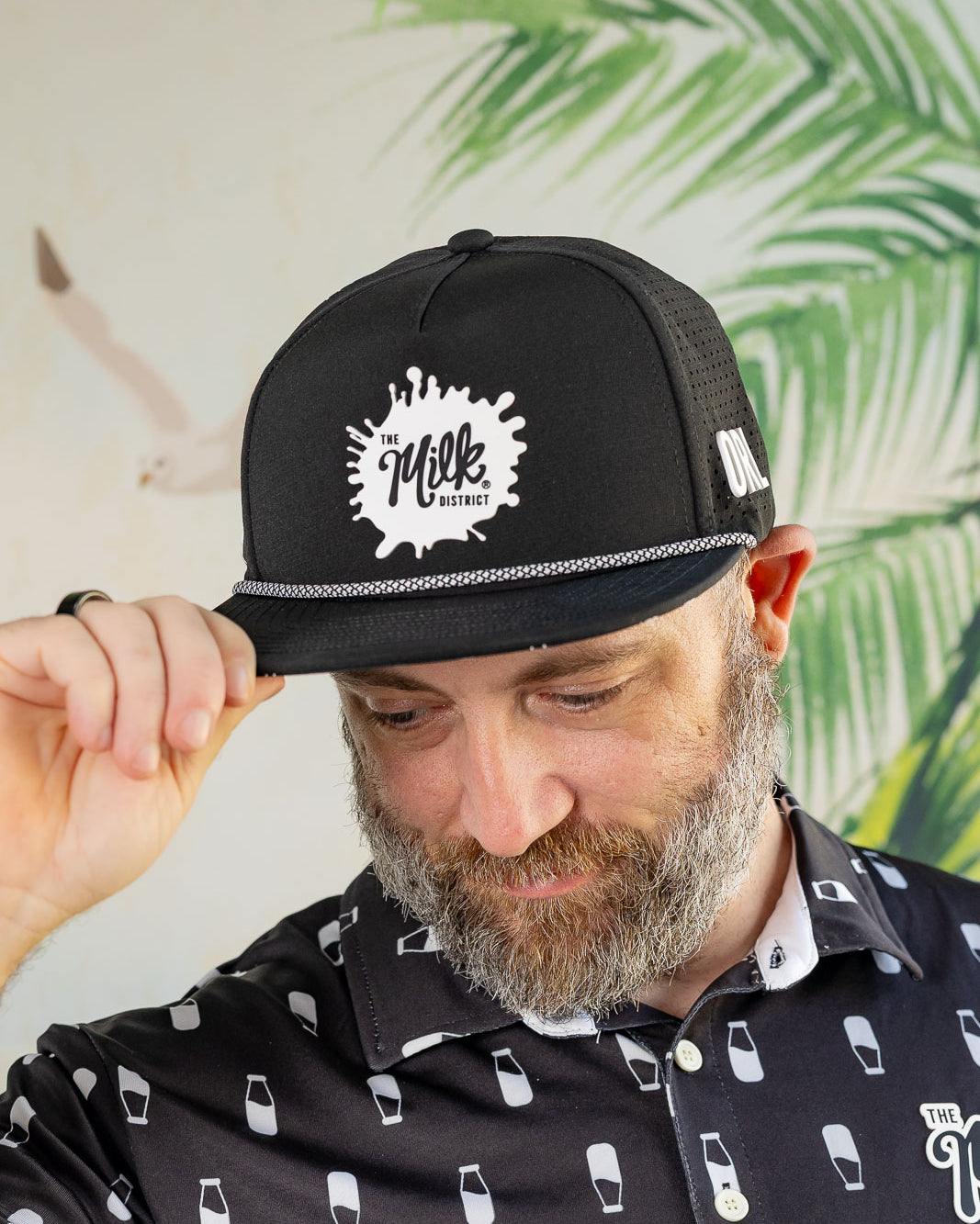 Man wearing a black cap with a logo and a patterned shirt against a leafy background