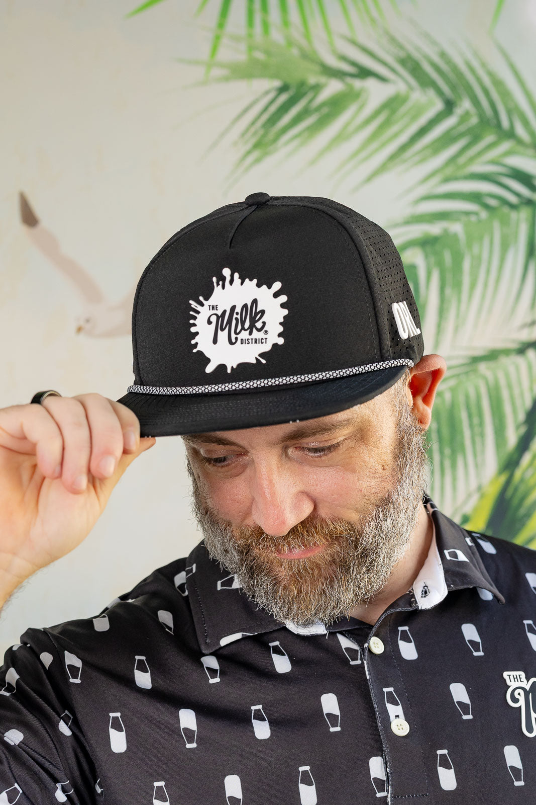 Man wearing a black cap with a logo and a patterned shirt against a leafy background
