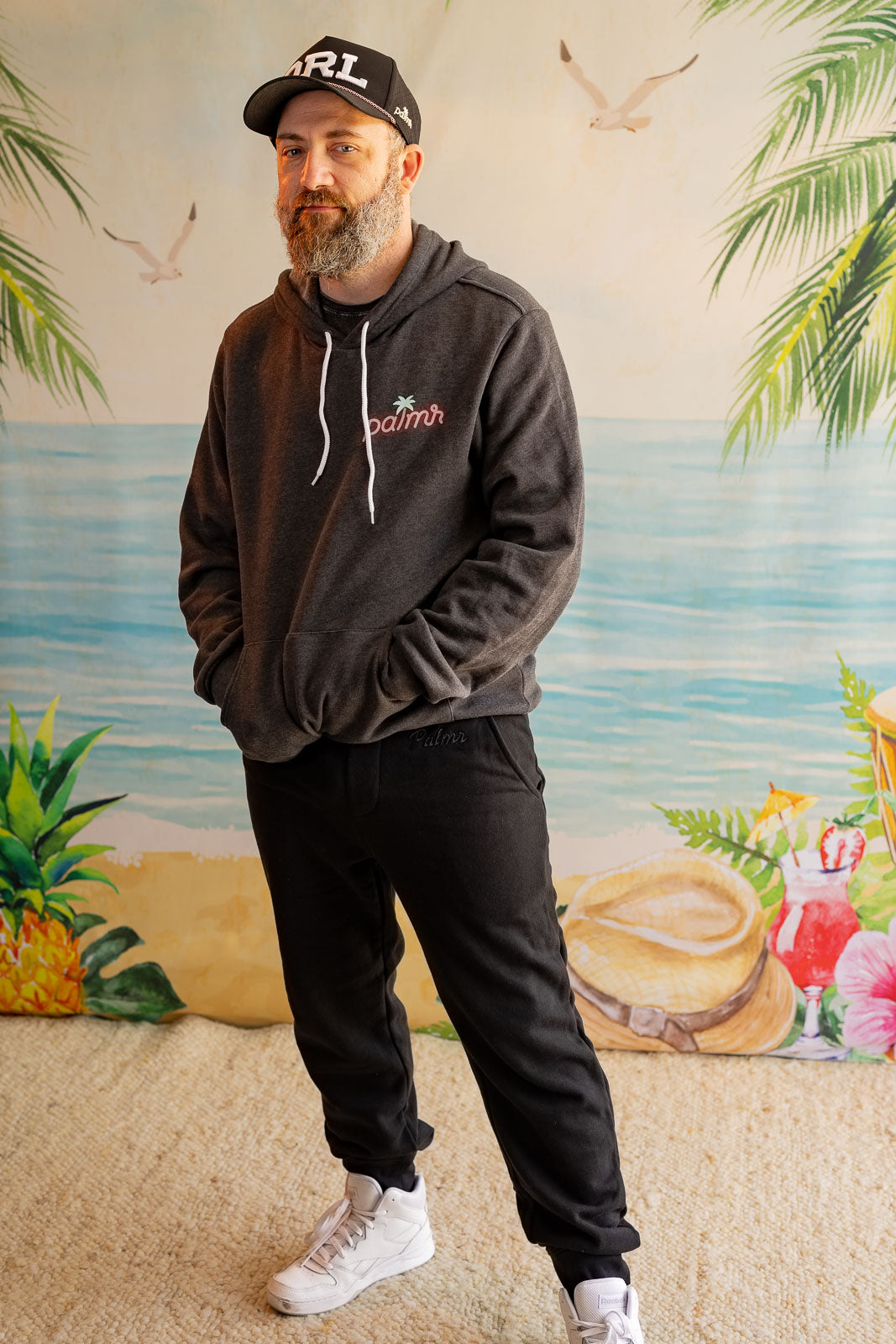 Man wearing a dark hoodie with a logo, black cap, and white sneakers standing in front of a colorful mural.