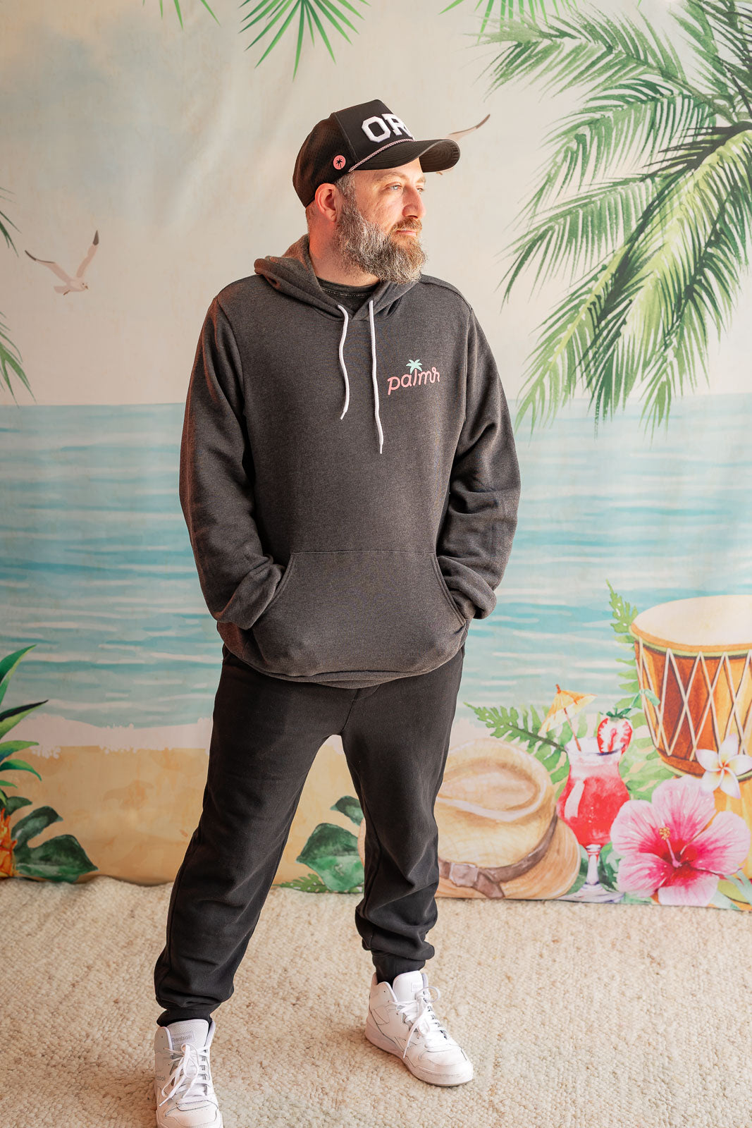 Man wearing a dark gray hoodie and pants with a logo, standing in front of a tropical-themed wall.