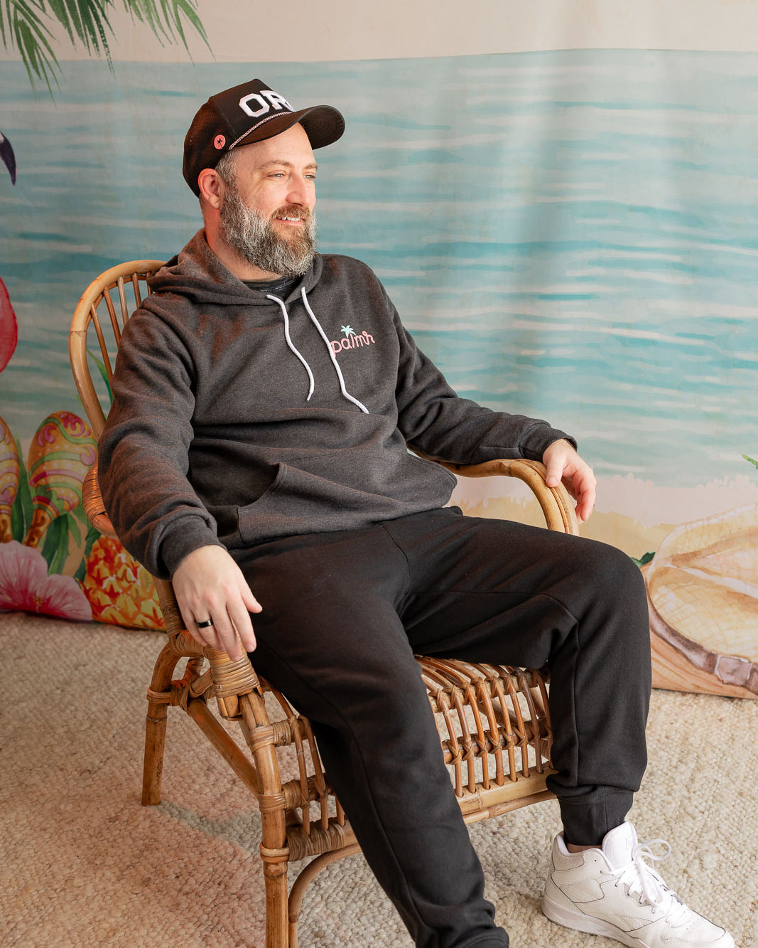 Man sitting on a wicker chair wearing a hoodie with a logo, in a room with a decorative wall.