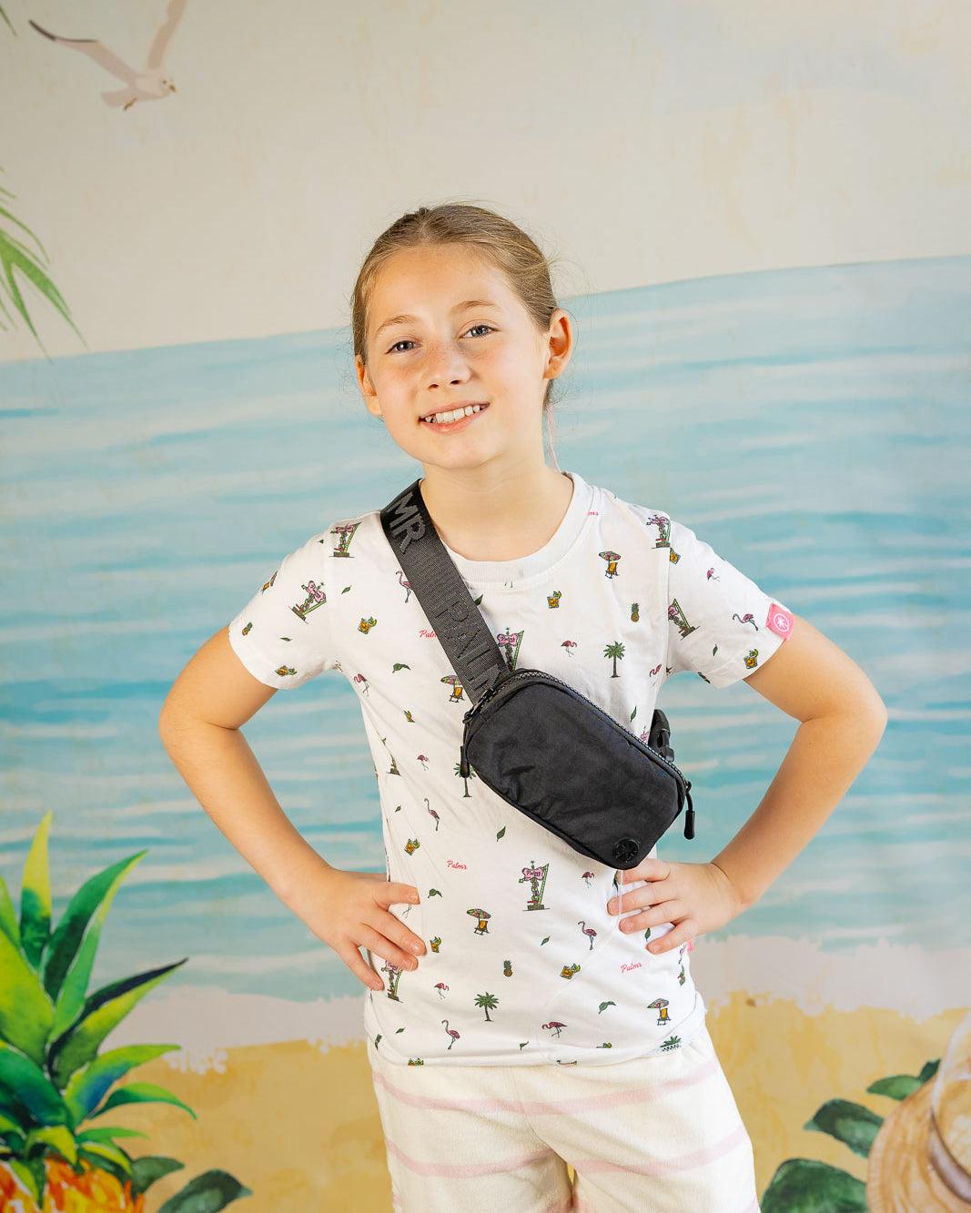 Child wearing a patterned shirt and shorts with a black waist bag, standing in front of a beach-themed wall.
