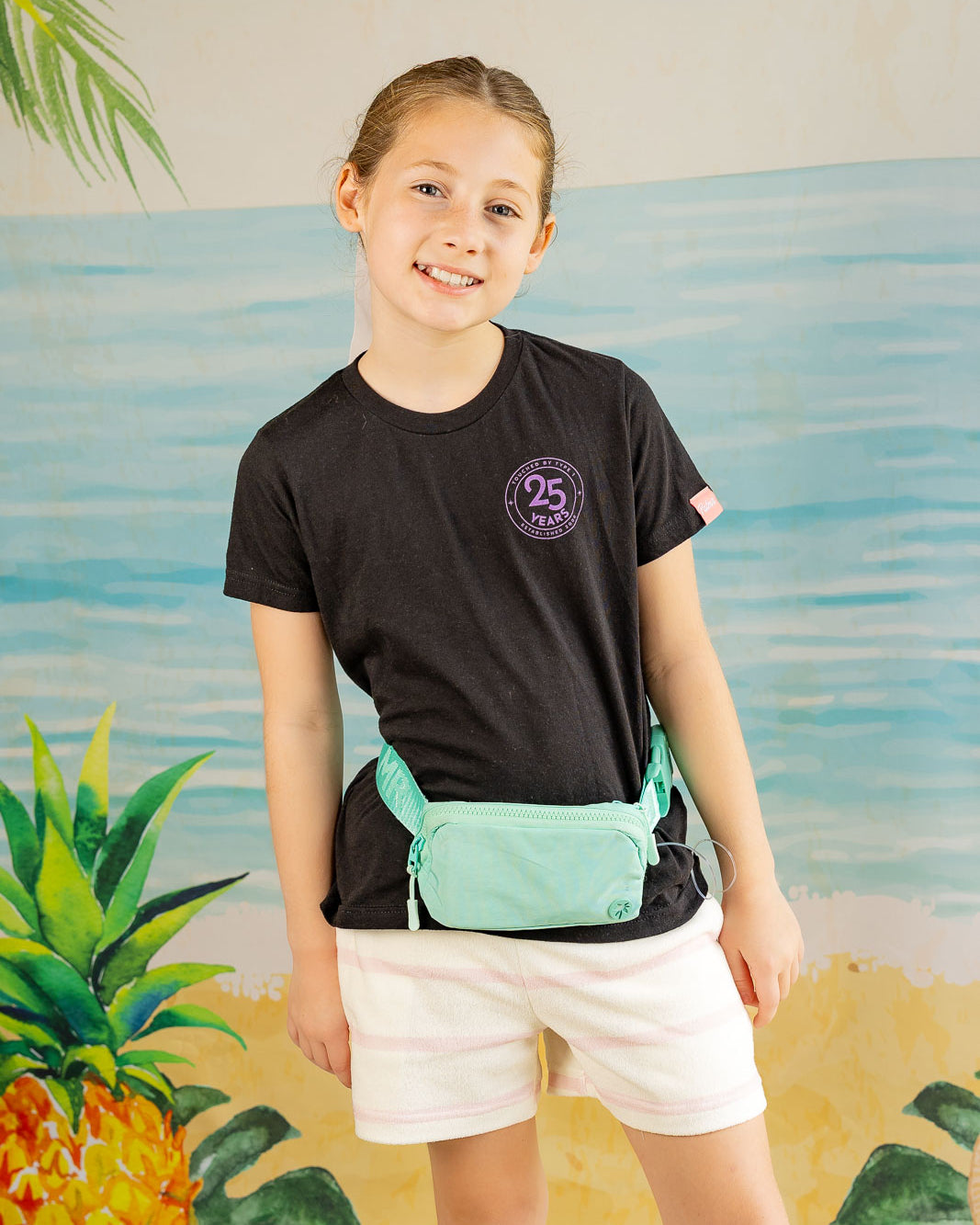 Child wearing a black t-shirt with a logo and white shorts, standing in front of a colorful mural.