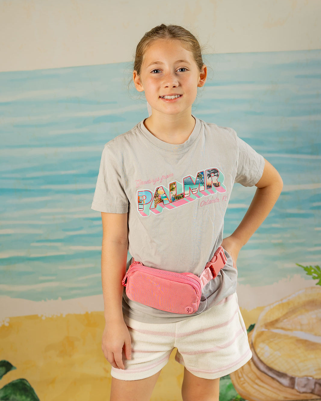 Young girl wearing a gray t-shirt with 'Palm' text and a pink fanny pack, standing in front of a beach-themed backdrop.