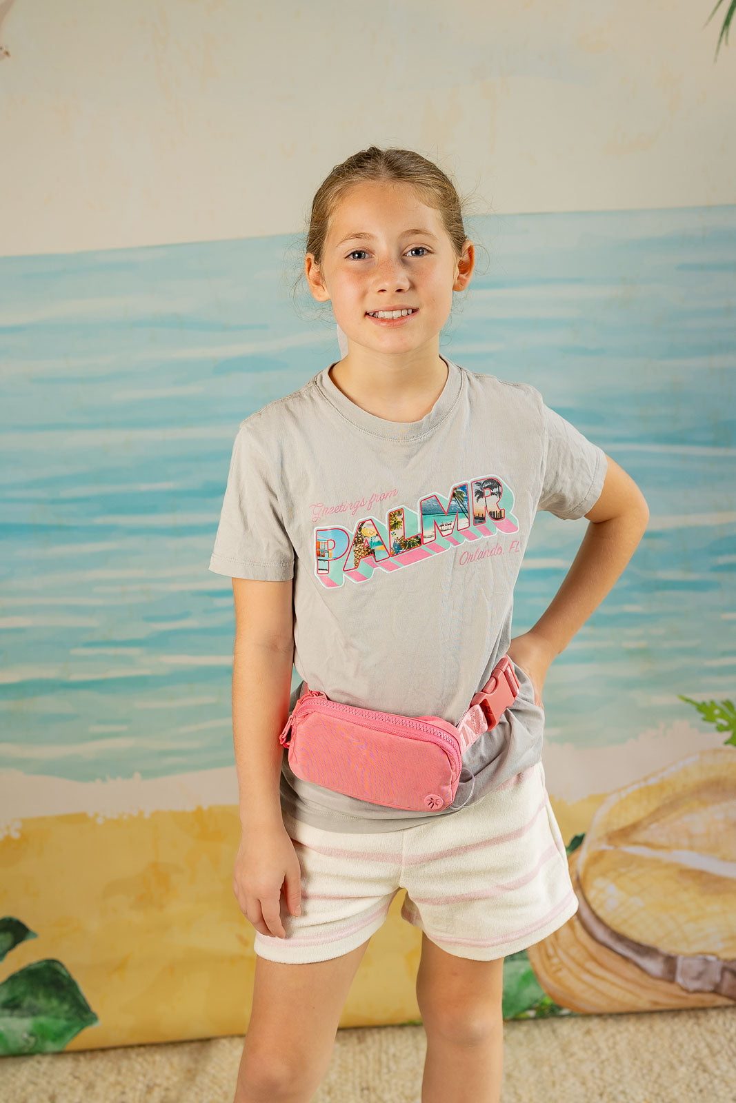 Young girl wearing a gray t-shirt with 'Palm' text and a pink fanny pack, standing in front of a beach-themed backdrop.