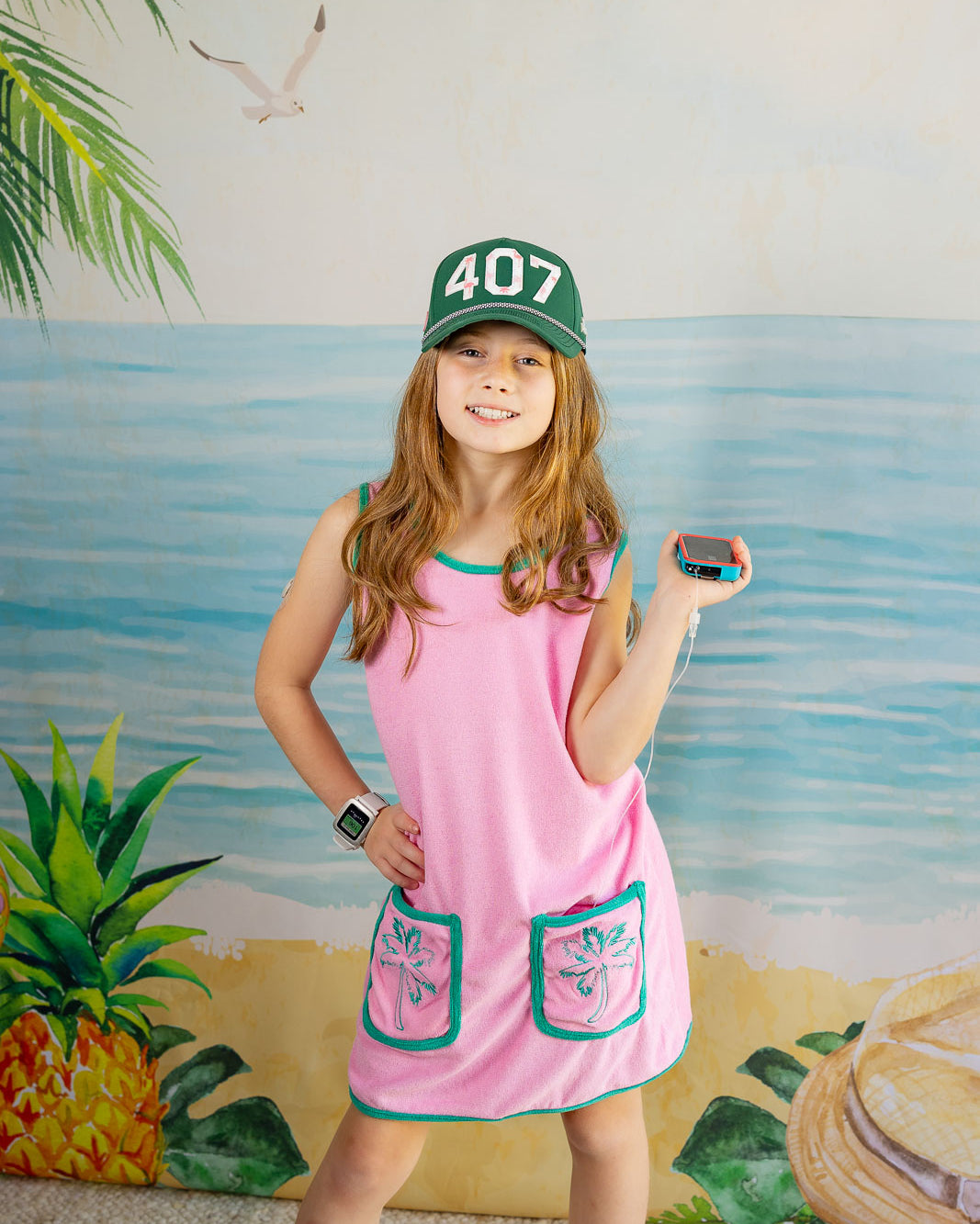 Young girl in a pink dress with green accents standing in front of a beach-themed wall.