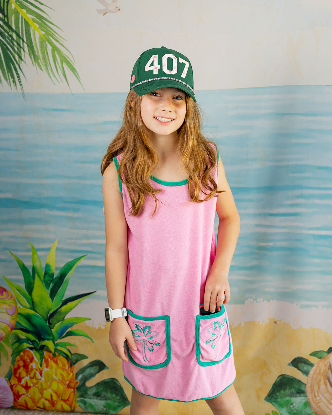 Young girl wearing a pink dress with green accents and a green cap with '407' on it, standing in front of a colorful mural.