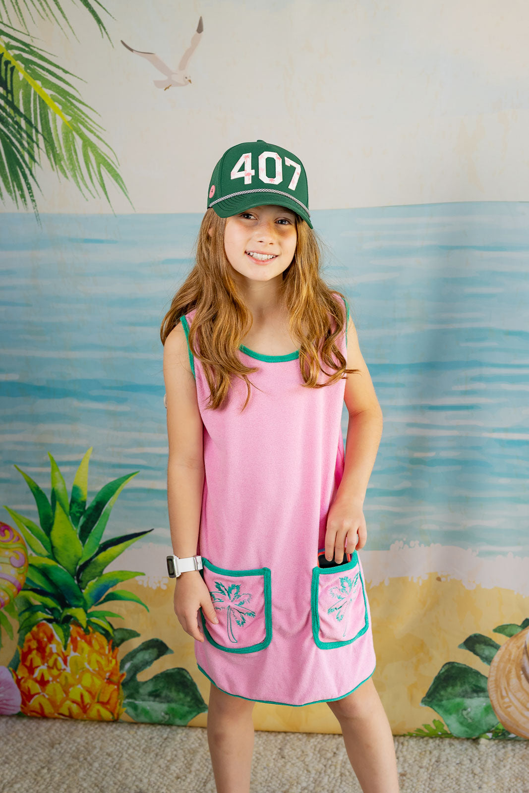 Young girl wearing a pink dress with green accents and a green cap with '407' on it, standing in front of a colorful mural.