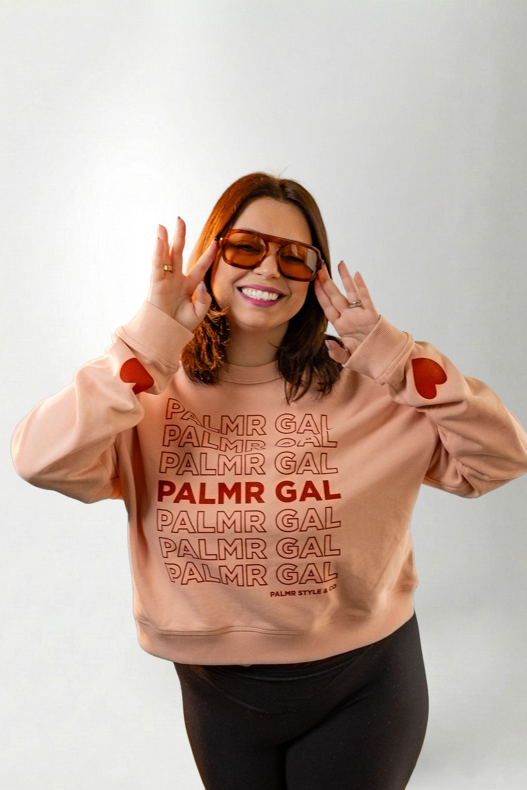 Person wearing a peach sweatshirt with 'PALMR GAL' text and heart designs on a plain background