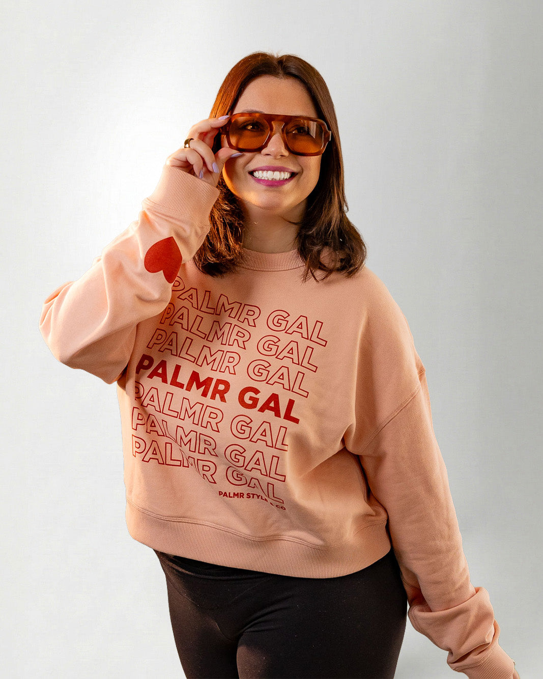 Person wearing a peach sweatshirt with 'PALMR GAL' text and heart designs on a plain background