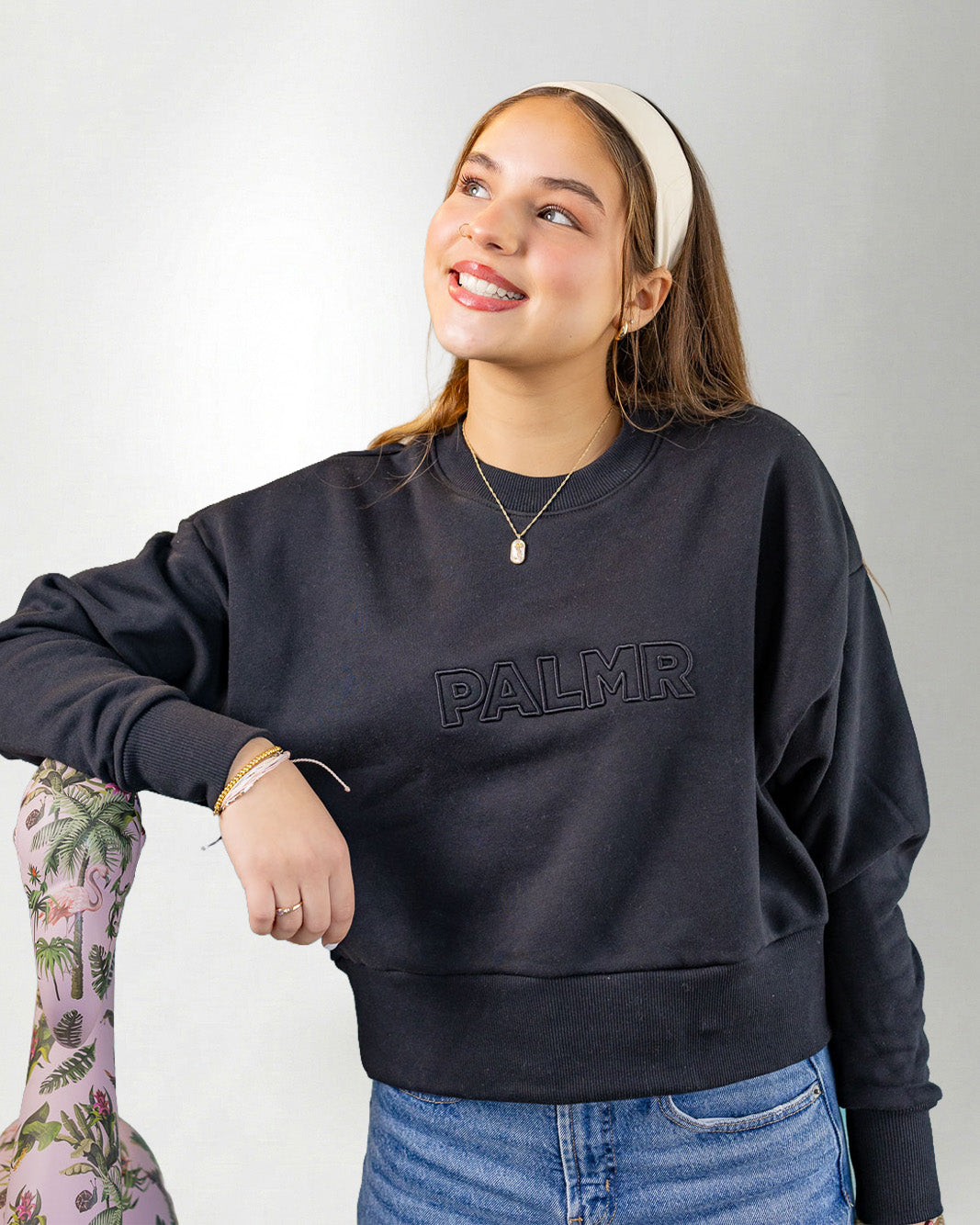 Woman wearing a navy blue sweatshirt with 'PALMR' branding, holding a floral-patterned bag against a white background.