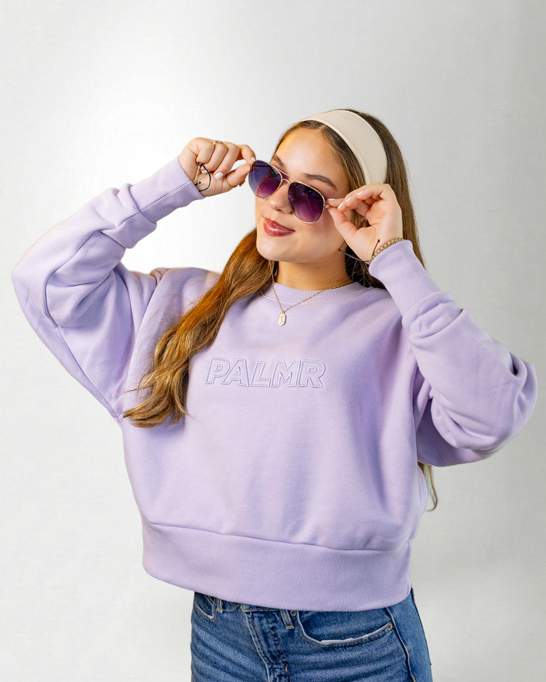 Woman wearing a lavender sweatshirt with 'PALM' branding, sunglasses, and a headband on a white background.