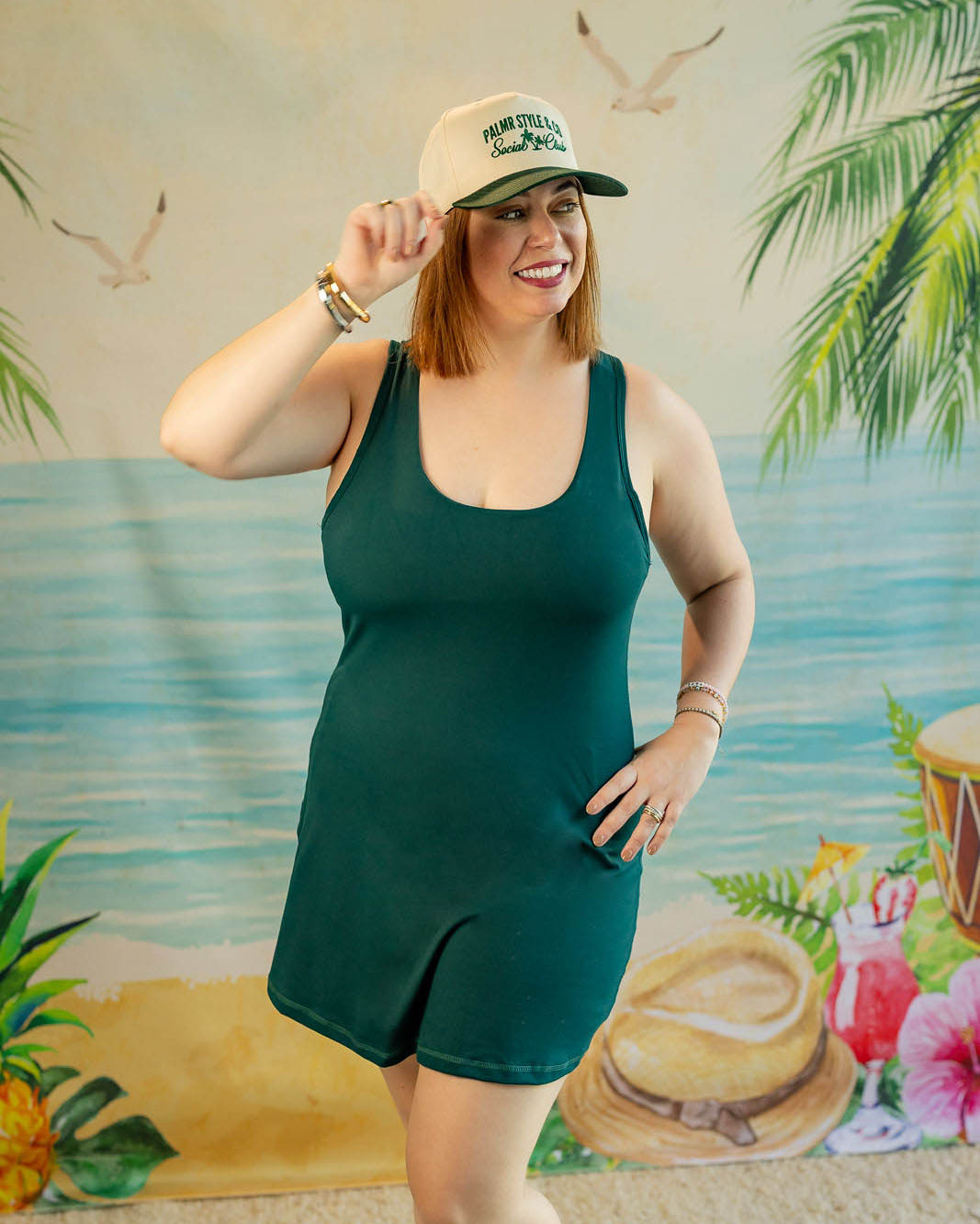 Woman in a green dress and cap posing in front of a tropical-themed backdrop