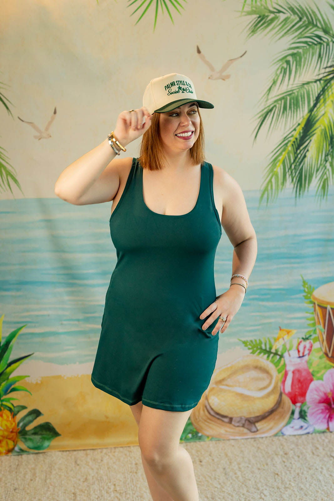 Woman in a green dress and cap posing in front of a tropical-themed backdrop