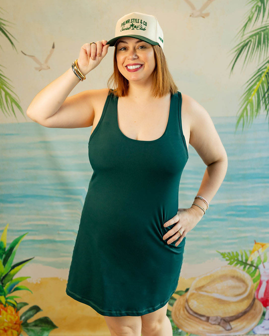 Woman wearing a green dress and white cap with palm tree design, standing in front of a tropical-themed backdrop.