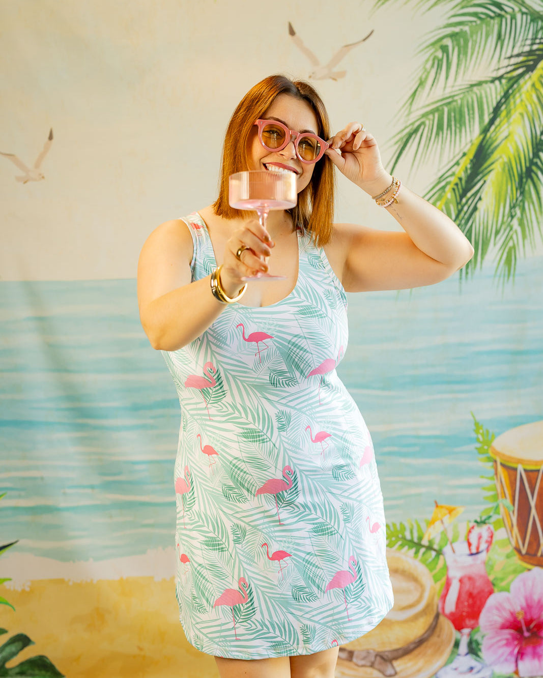 Woman in a floral dress holding a glass against a tropical-themed backdrop