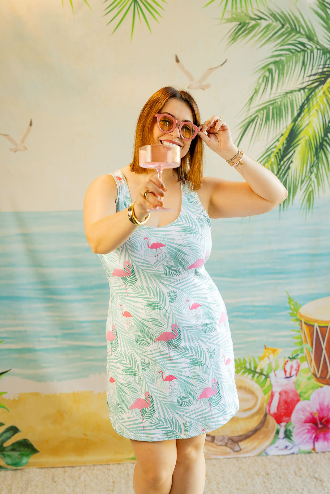 Woman in a floral dress holding a glass against a tropical-themed backdrop