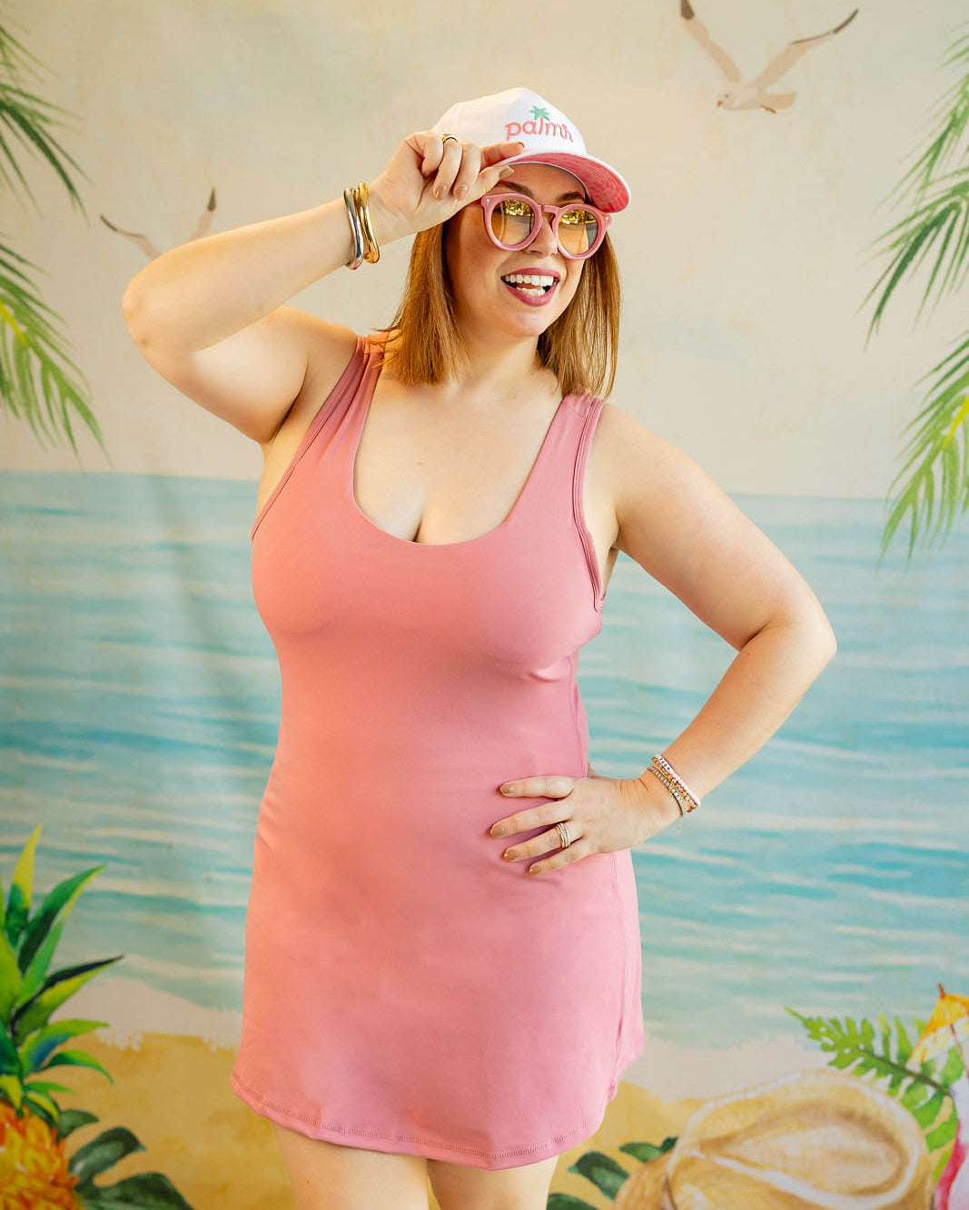 Woman in a pink dress and cap posing in front of a tropical-themed backdrop