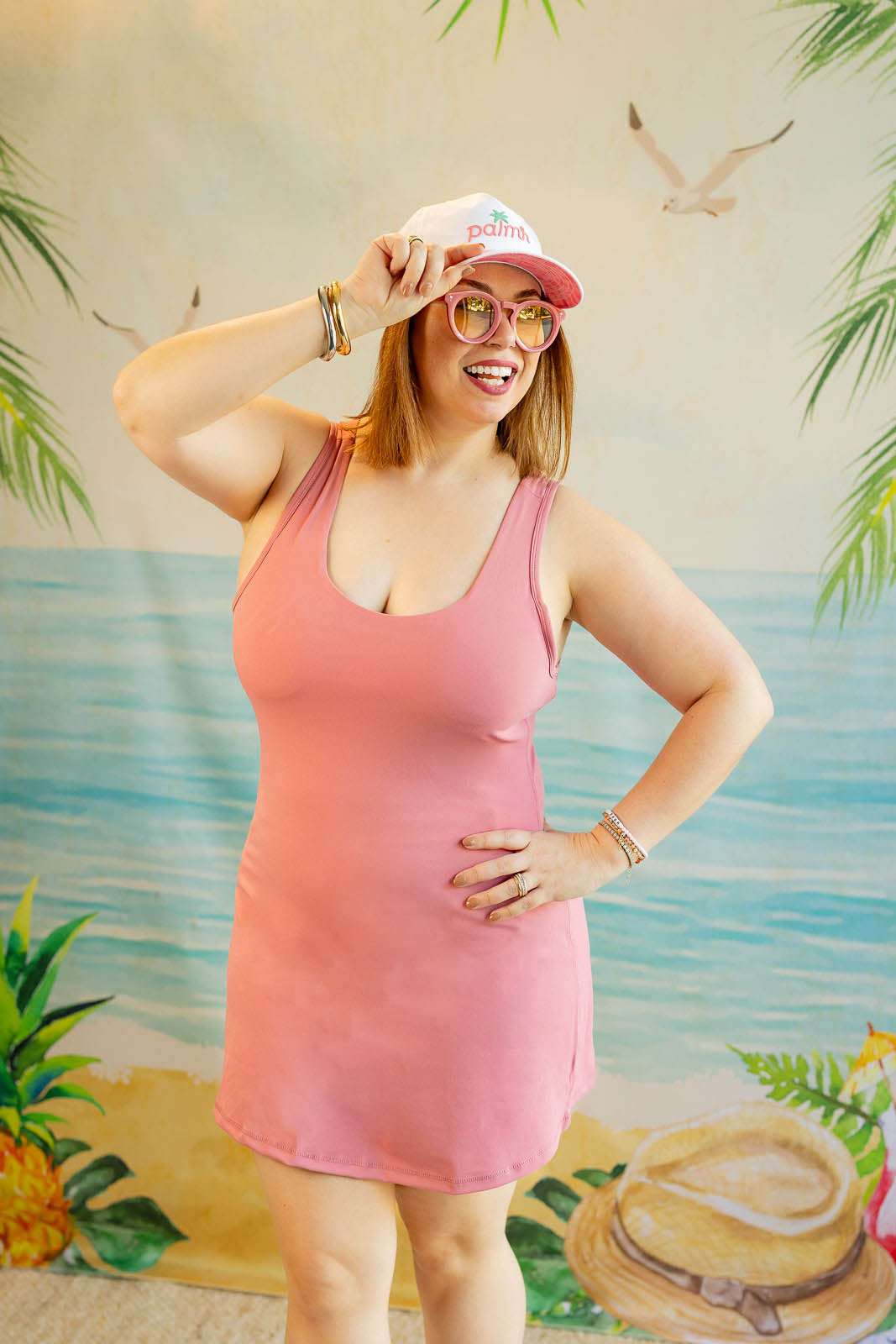 Woman in a pink dress and cap posing in front of a tropical-themed backdrop