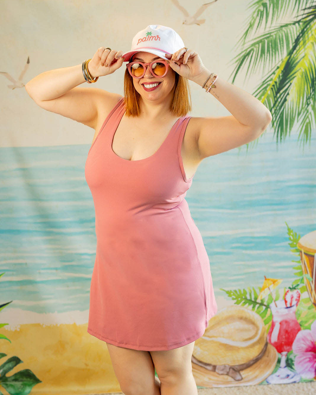 Woman in a pink dress and white cap posing in front of a tropical-themed backdrop