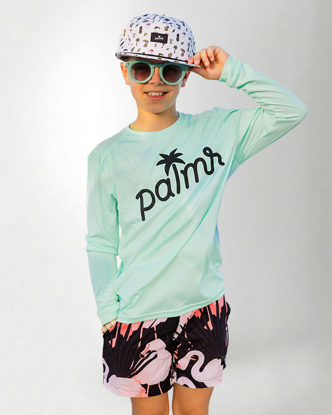 Person wearing a light green long-sleeve shirt with 'palms' logo, colorful cap, sunglasses, and flamingo shorts on a white background.