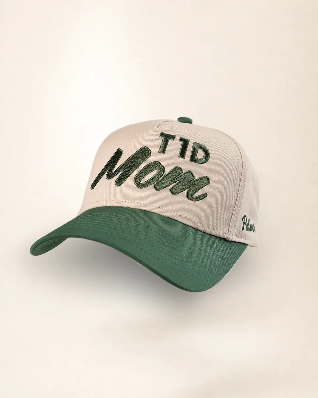 A baseball cap with a light beige crown and a dark green brim, featuring 'TID Mom' embroidered on the front in dark green script, with a small 'Palm.