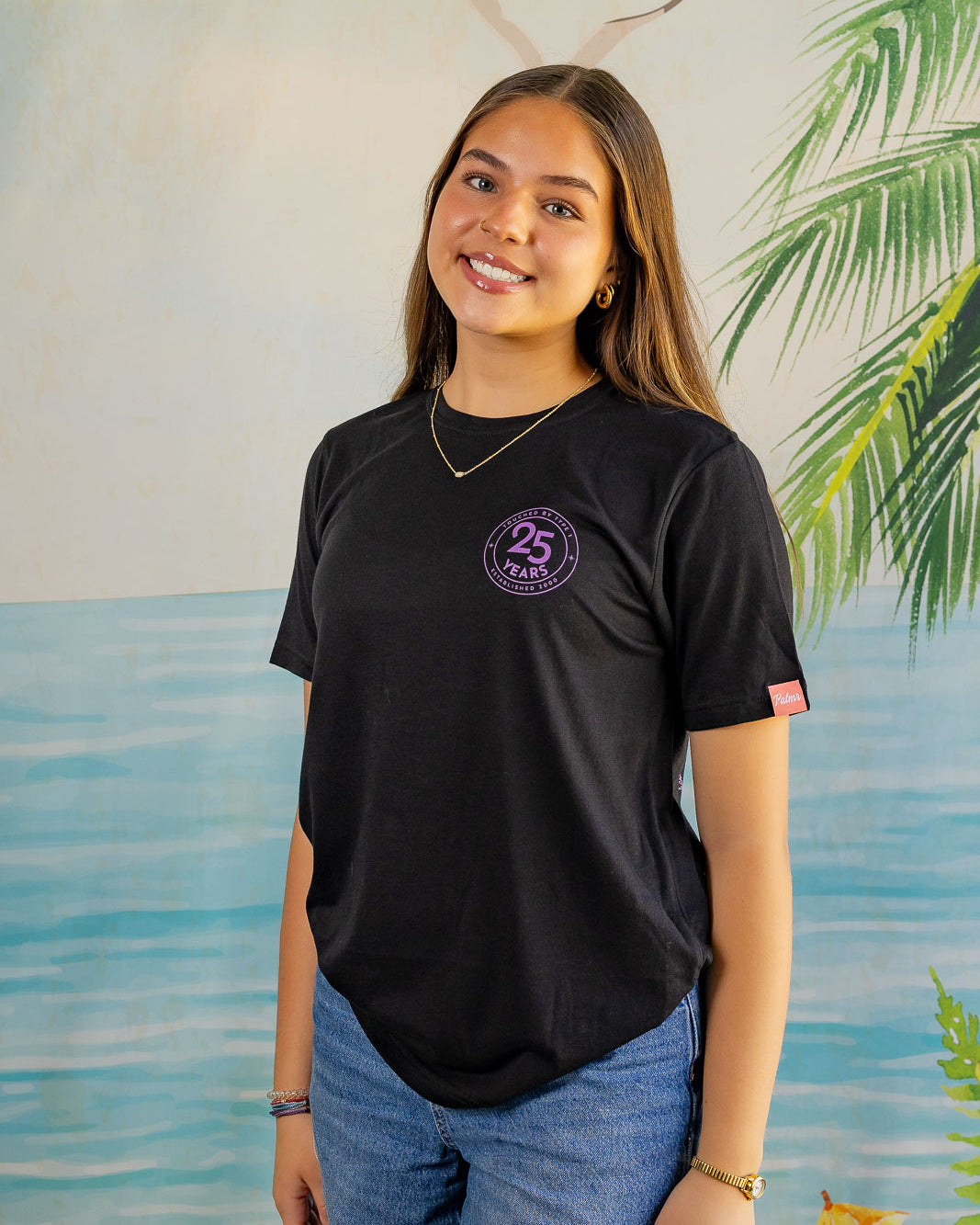 Person wearing a black t-shirt with a logo, standing in front of a scenic background with water and plants.